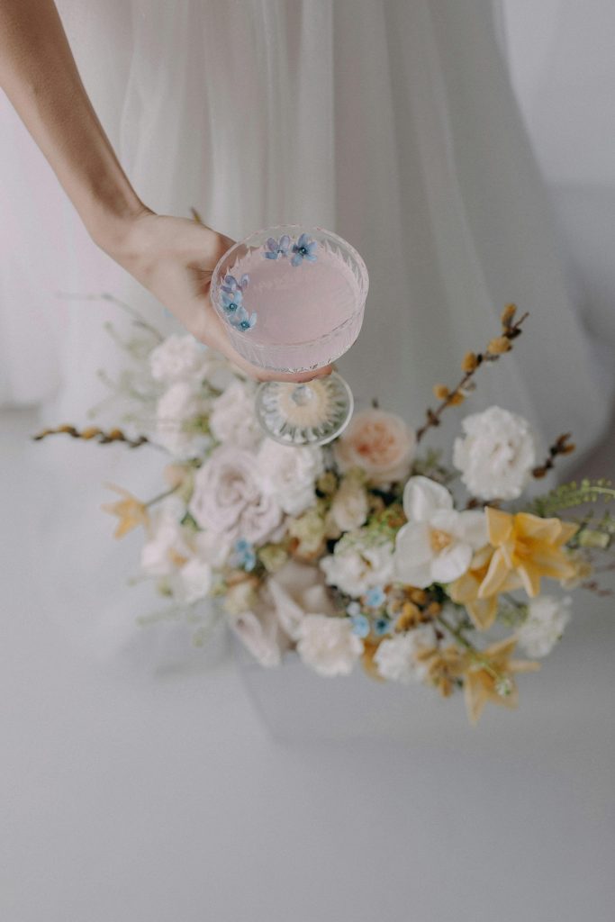Bride in white gown holding a decorated cocktail glass with a floral bouquet, showcasing wedding elegance.